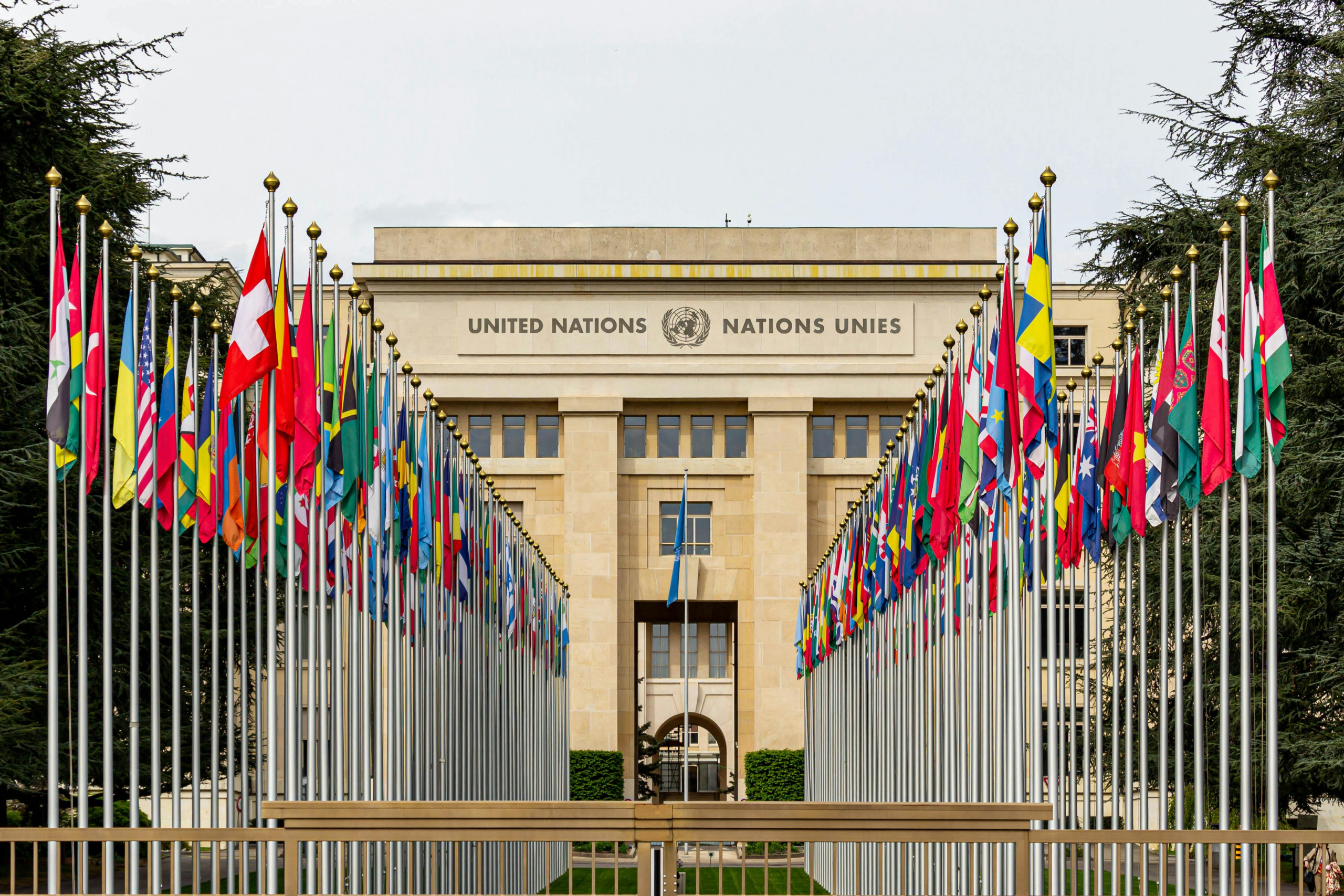 Front view of the United Nations Office in Geneva, with numerous international flags lined up on both sides leading to the building.