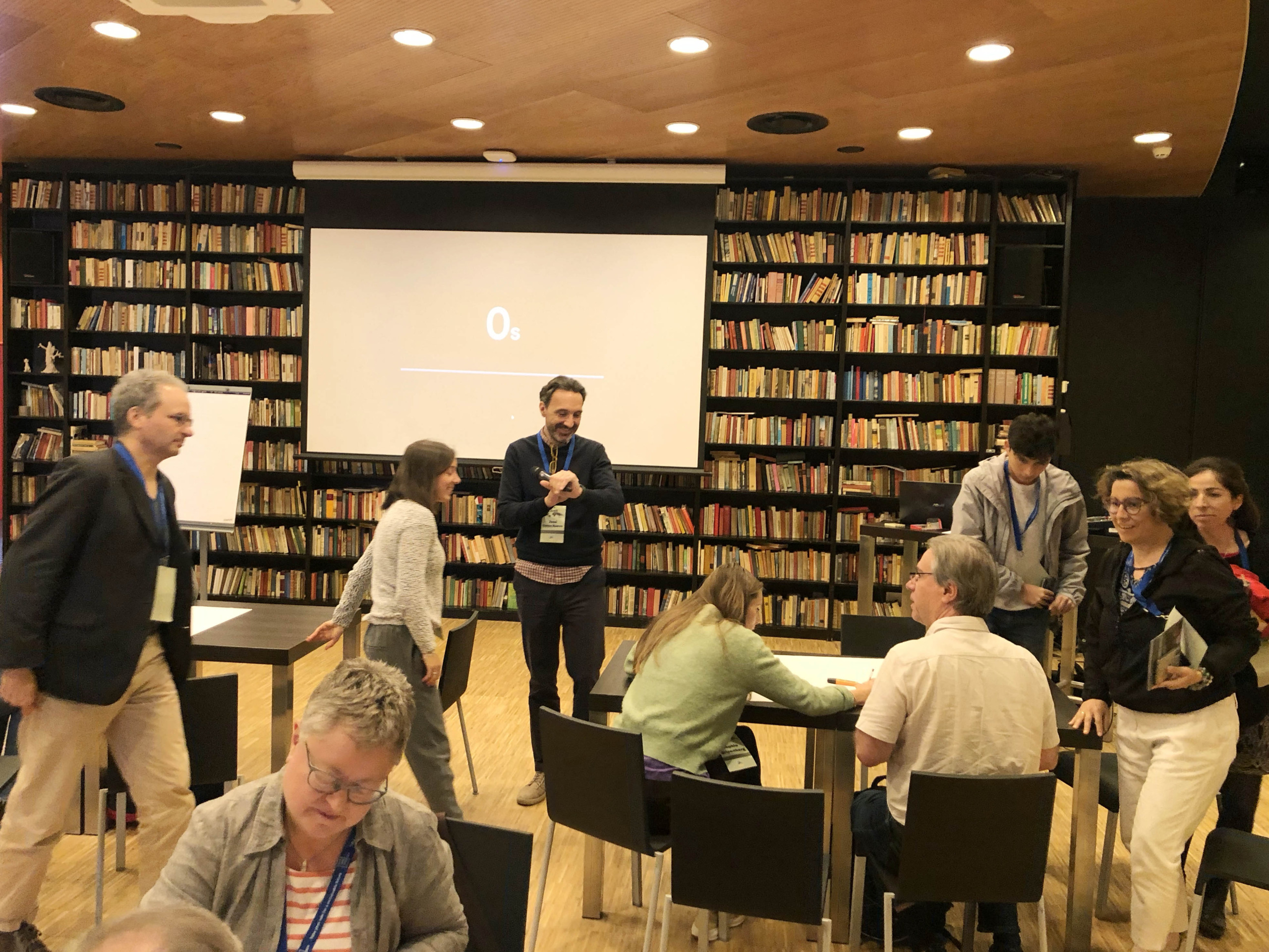 A group of people engaging in a discussion in a library setting, with bookshelves in the background and a projector screen displaying 0s. Some individuals are seated, while others stand or walk around.